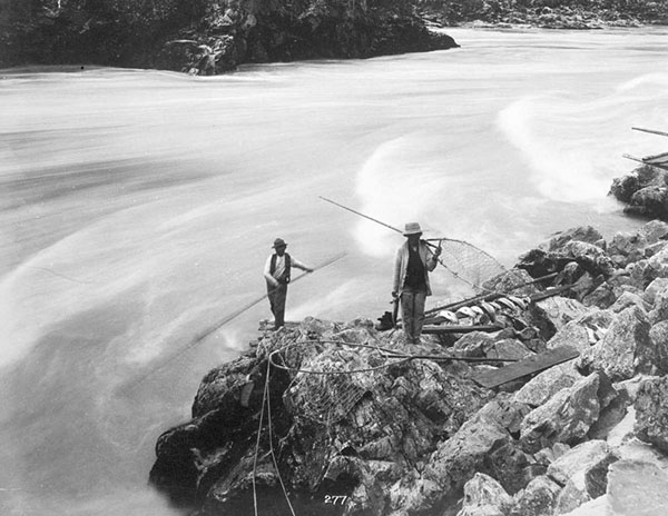 Two Stó꞉lō men fish using dipnets next to a rushing river.