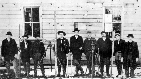 Group of ten men stand outside by a wooden wall with the surveying equipment, circa 1890s.