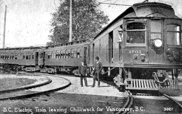 Two men stand next to the BC Electric Train engine with passenger cars leaving for Vancouver from Chilliwack, circa 1910.