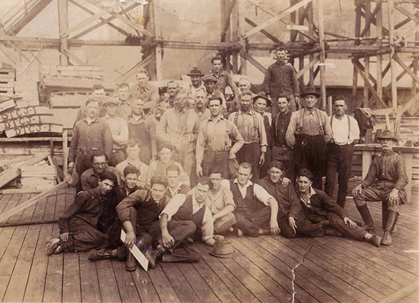 Group of millworkers gathered in front of wooden scaffolding circa 1912.