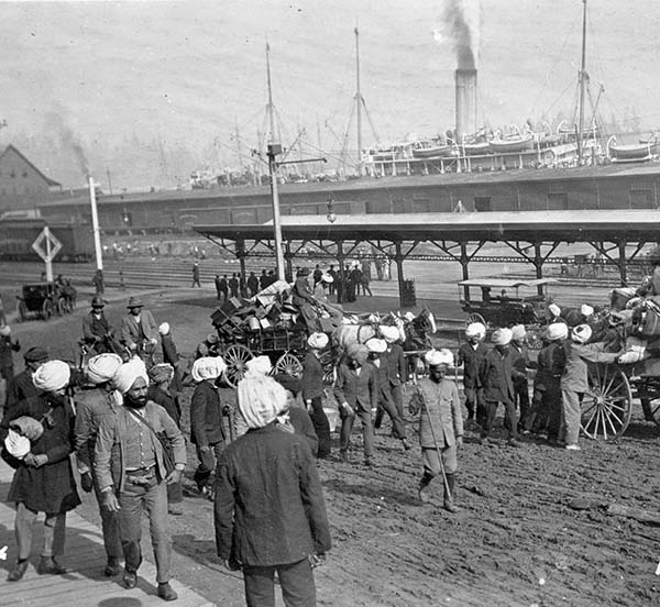 Group of men in turbans stand in front of a ship at anchor.