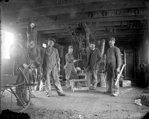 Group of men and a horse pose with tools in a blacksmith shop.