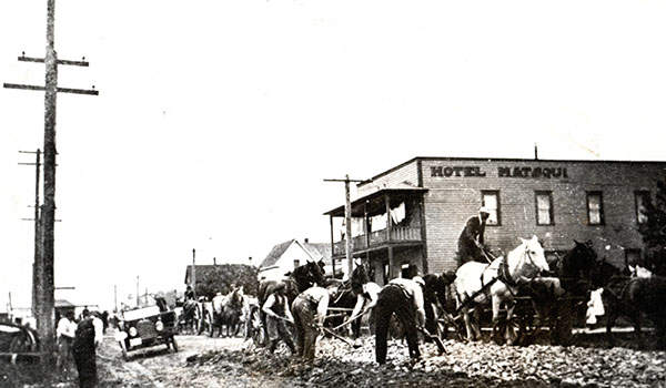 Group of men bent over with pickaxes working on the road in front of a large porched building with the sign "Hotel Matsqui".