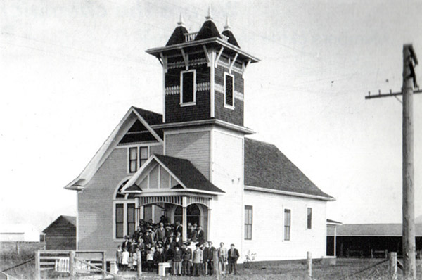 Group of people stand on the front steps of a timber-framed church with patterned shingles and steeple with four spires on the roof.