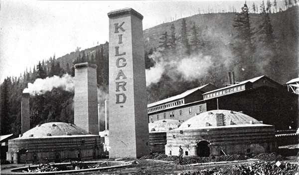 Several smokestacks, including one with a sign reading "Kilgard", in front of a factory next to concrete domed brick kilns.